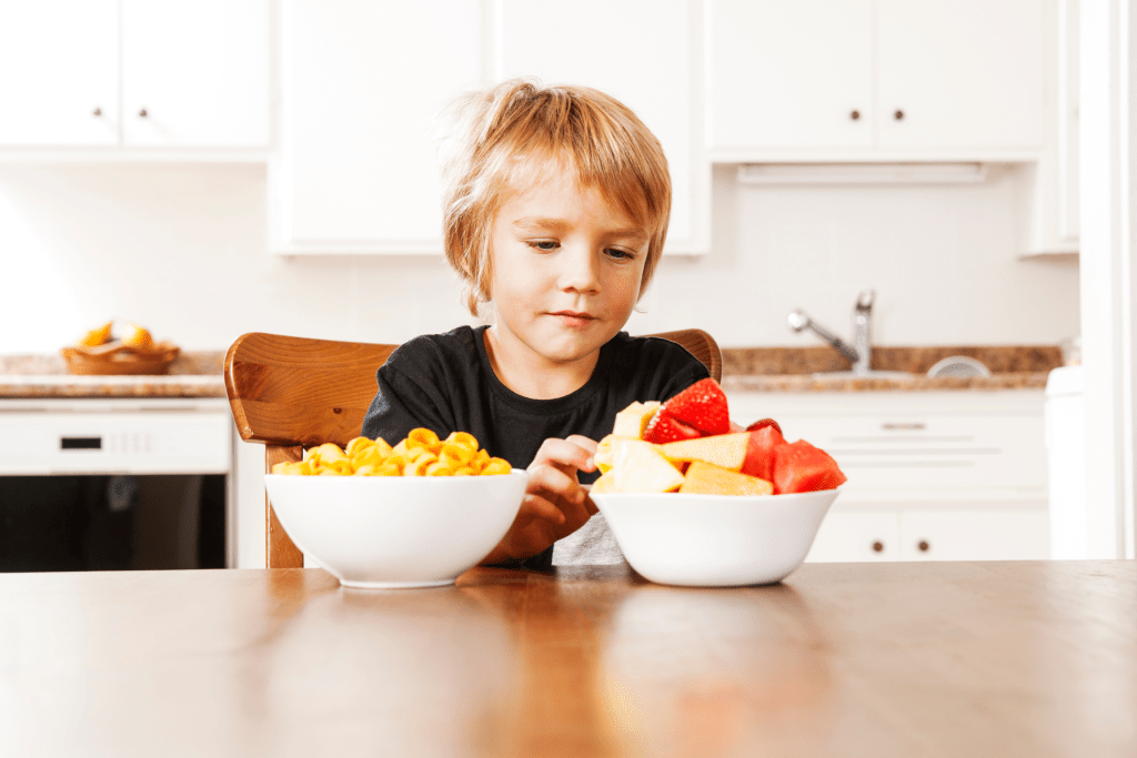 A young boy sitting at a table, thoughtfully choosing between a bowl of fruit and a bowl of snacks, fostering decision-making skills. 