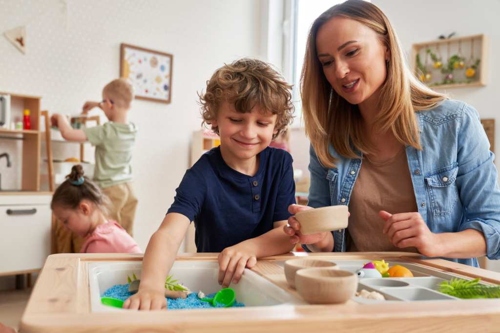 Smiling boy exploring a sensory bin with a therapist during an autism-focused play session in a classroom during summer