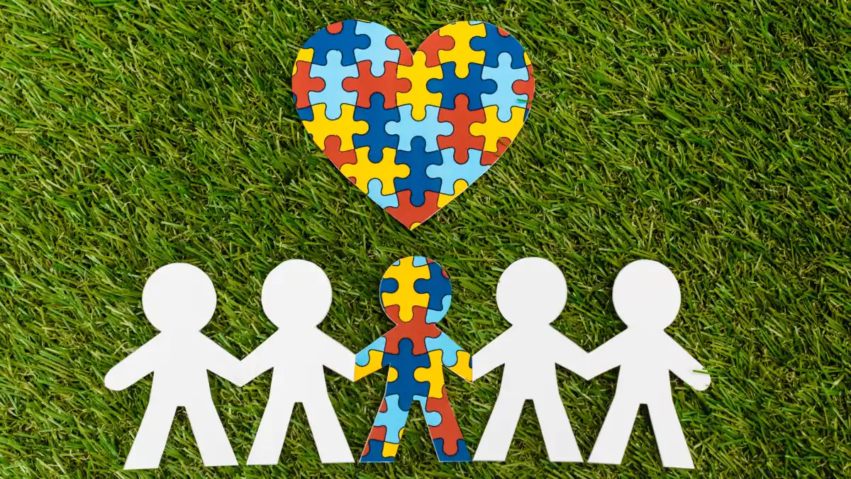 Paper chain of five people, the one in the middle is colorful with the autism puzzle pieces and a similar heart above. The other four people, two on each side, are plain white.