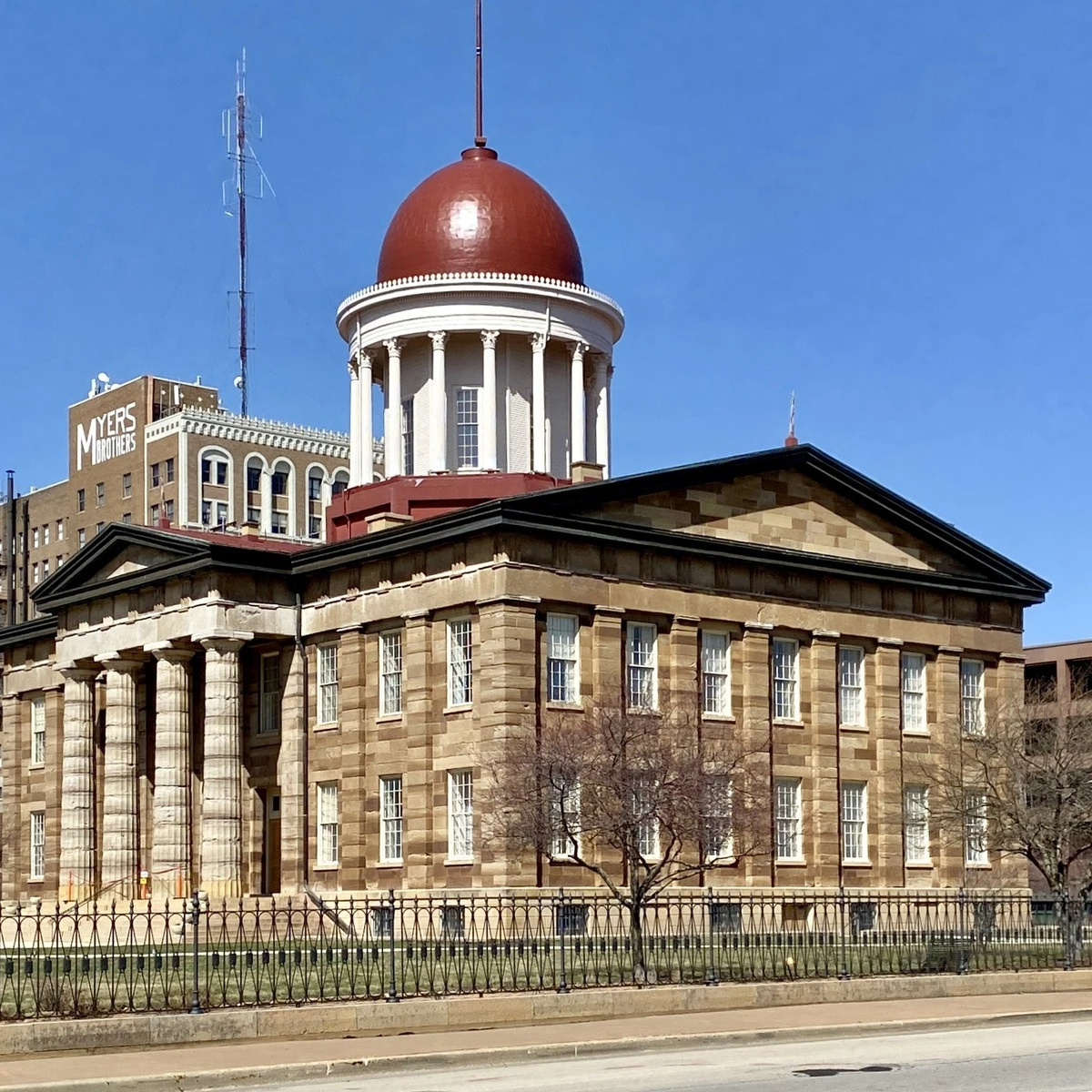 Historic brick government building with a white-columned rotunda and red dome, set behind a wrought-iron fence under a clear blue sky.