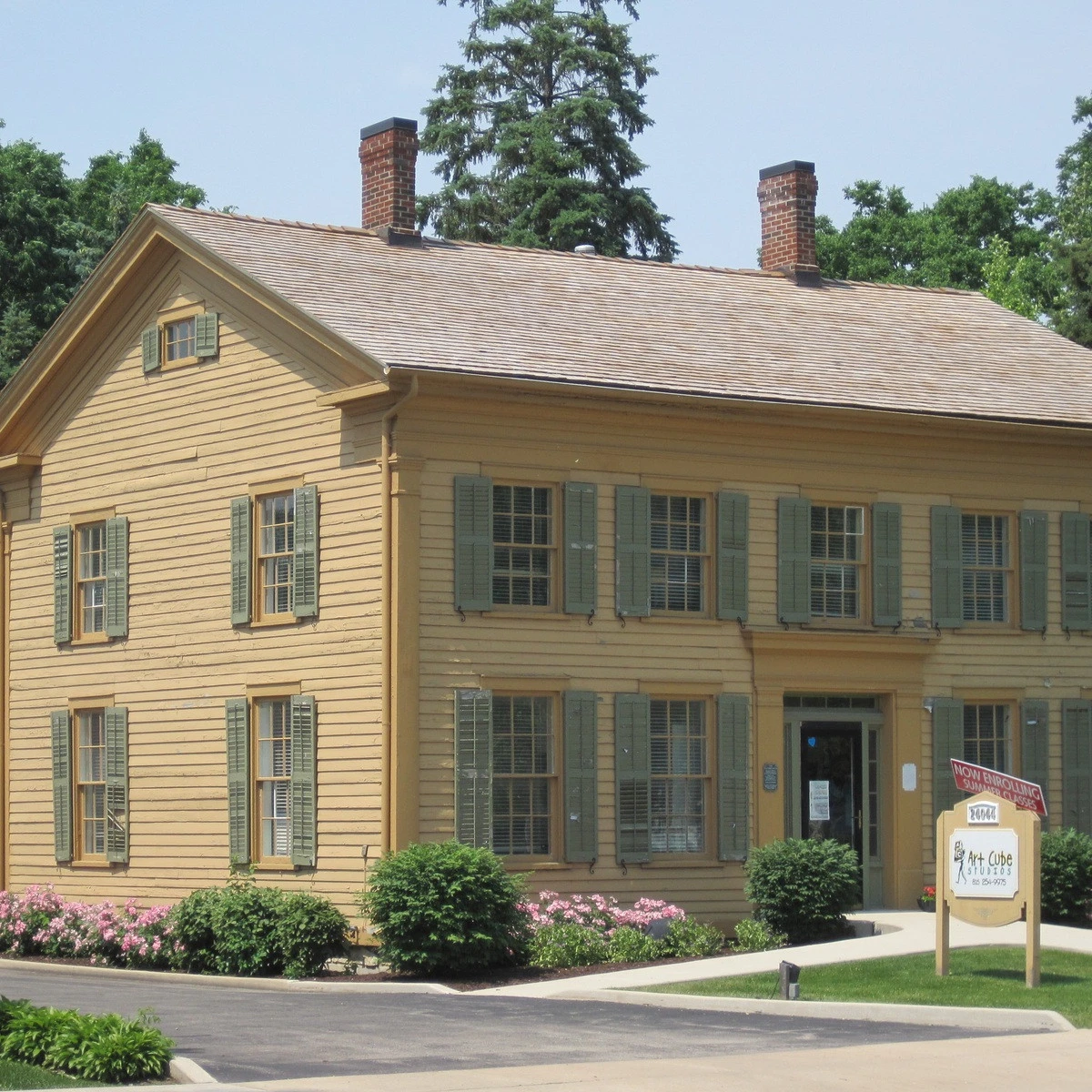 Two-story yellow clapboard building with green shutters and a small garden in front near a sidewalk signboard outside the entrance.