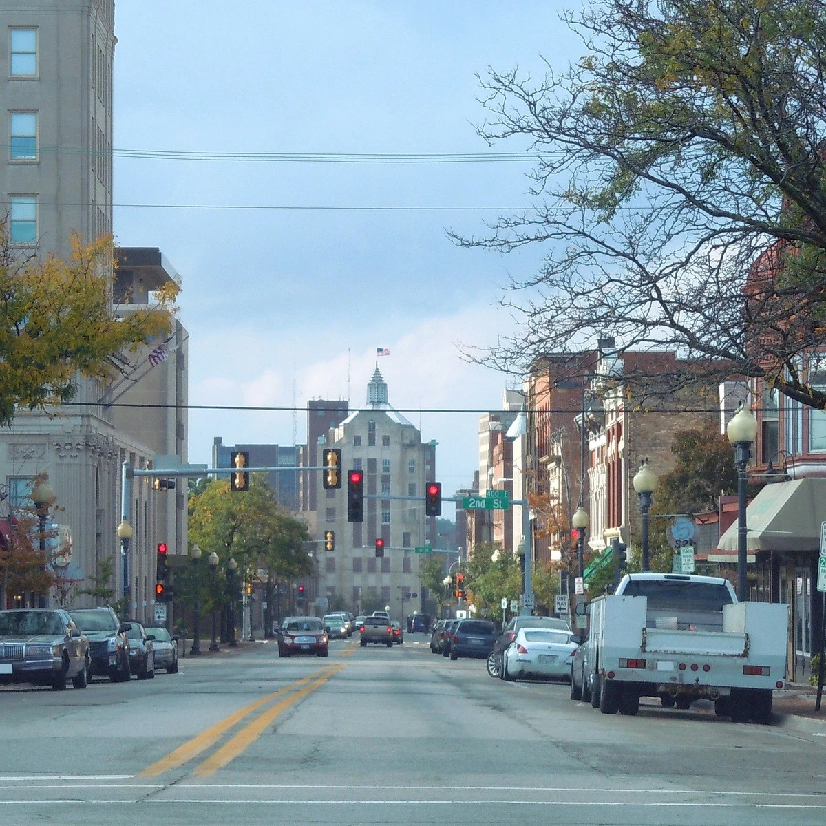 Urban street view with cars parked along both sides and a distant, ornate city hall-style building at the end of the block under a blue sky with scattered clouds.